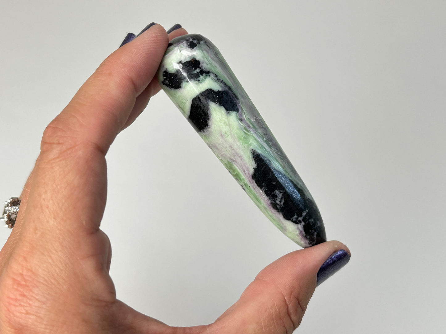 A hand with dark blue nail polish holds a polished, teardrop-shaped Rare Green and Purple Kammererite Wand showing a marbled pattern of black, light green, white, and subtle lavender veins against a plain light gray background.