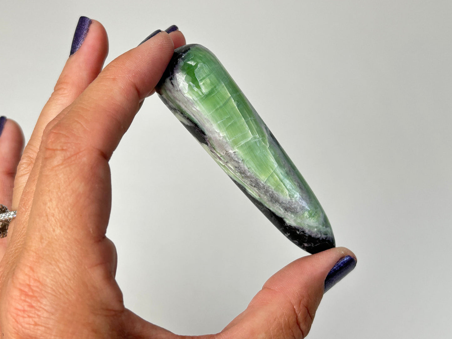 A hand with dark purple nail polish holds a polished Rare Green and Purple Kammererite Wand with prominent white and black banding, set against a plain light gray background.