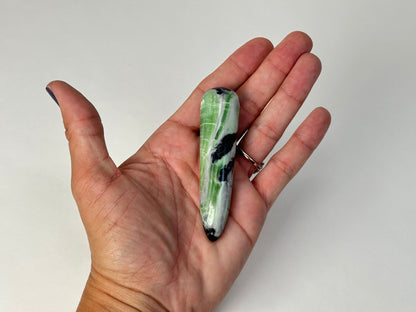A hand holding a polished Rare Green and Purple Kammererite Wand against a plain light gray background.