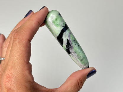 A hand with dark blue nail polish holds a polished Rare Green and Purple Kammererite Wand against a plain light gray background.
