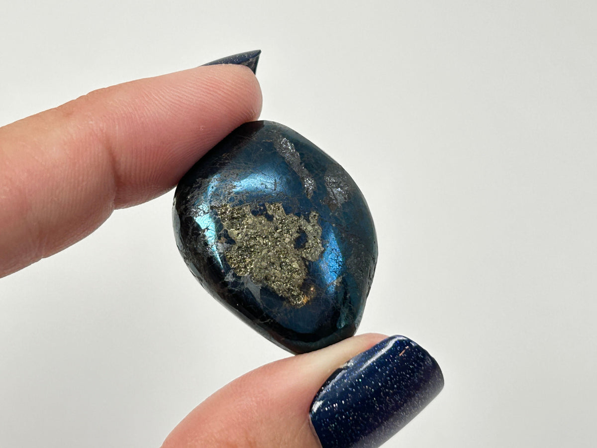 A polished dark blue Rare Healing Covellite Stone with a metallic gold inlay, held between two fingers with dark glittery nail polish against a plain light gray background.