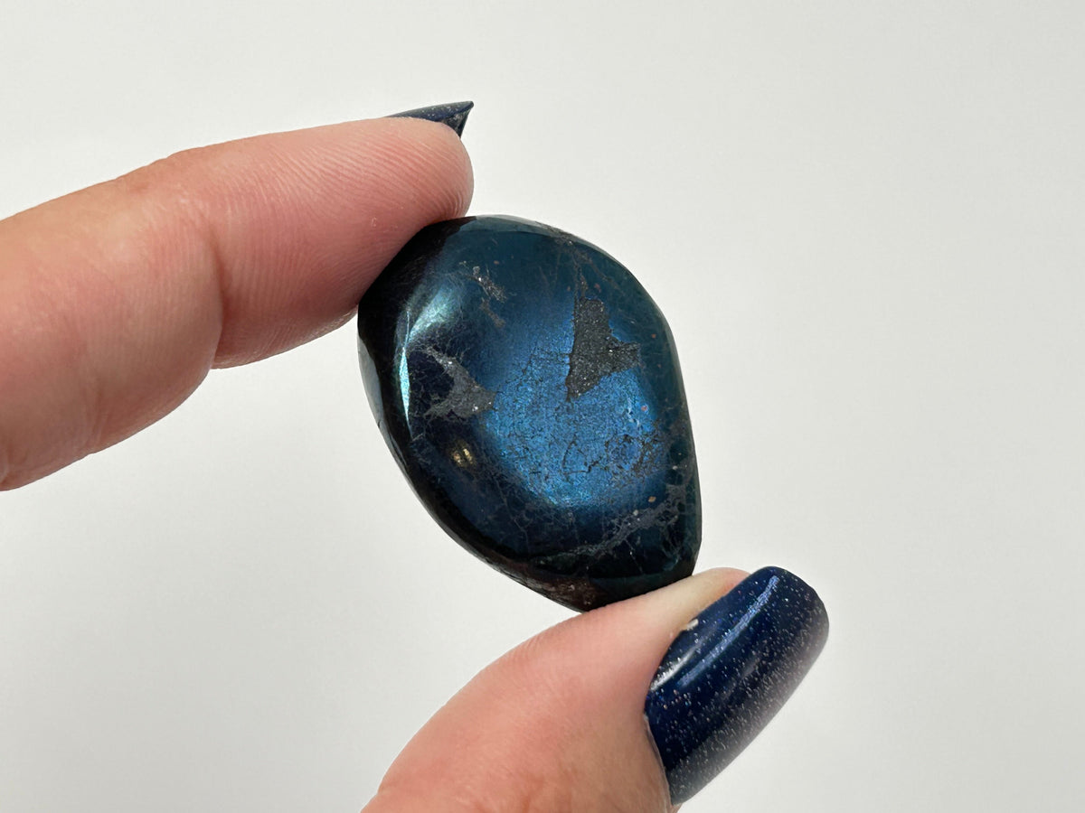 A polished dark blue Rare Healing Covellite Stone held between two fingers with blue glitter nail polish, against a plain white background.