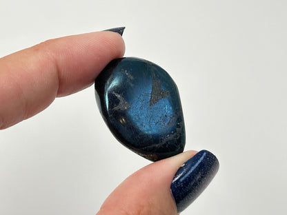 A polished dark blue Rare Healing Covellite Stone held between two fingers with blue glitter nail polish, against a plain white background.