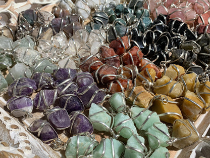 A close-up display of multiple raw stone pendants in silver wire settings, arranged in rows. The stones include purple amethyst, green aventurine, yellow howlite, light green malachite, clear quartz, dark gray, pink, and reddish brown, set against a light wood background.