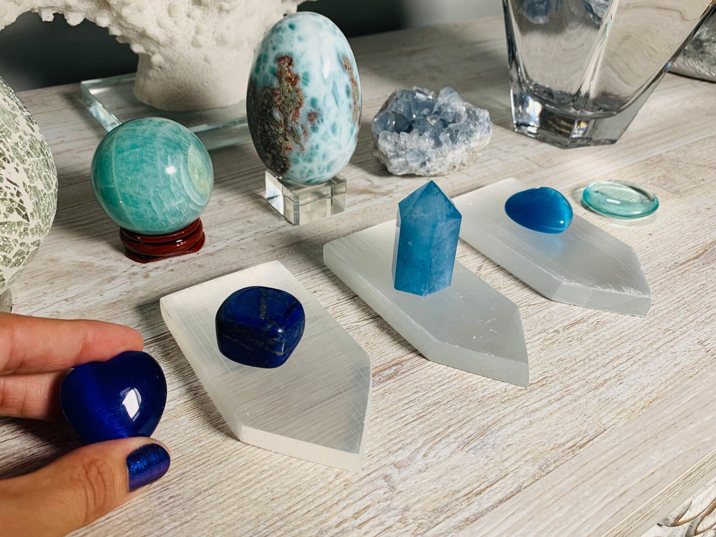 A collection of blue and turquoise crystals displayed on white geometric stands on a light wood table, with a hand holding a polished deep blue stone in the foreground.