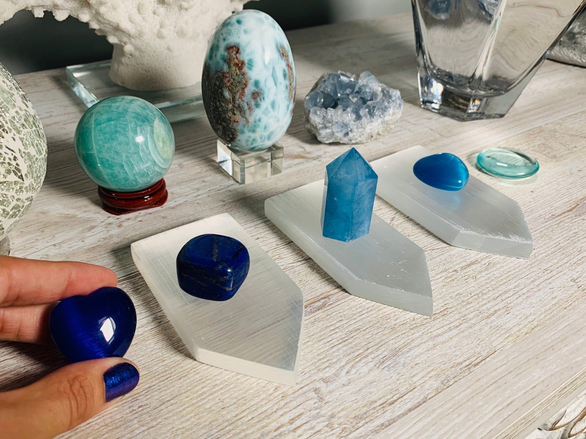 A collection of blue and turquoise crystals displayed on white geometric stands on a light wood table, with a hand holding a polished deep blue stone in the foreground.