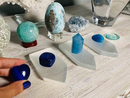 A collection of blue and turquoise crystals displayed on white geometric stands on a light wood table, with a hand holding a polished deep blue stone in the foreground.