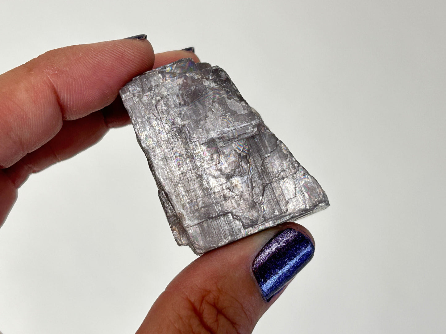 A hand holding a rough, metallic silver-gray Soothing Purple Anhydrite Stone specimen against a plain light background.