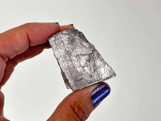 A hand holding a rough, metallic silver Soothing Purple Anhydrite Stone with a textured, fibrous crystalline structure against a plain white background.