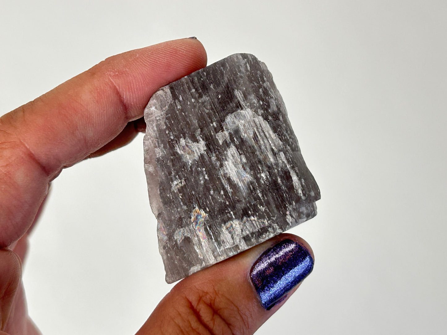 A hand holding a rough, polished gray Soothing Purple Anhydrite Stone against a plain light background. The person's thumb and index finger are visible, with the fingernail painted in a metallic purple-blue polish.