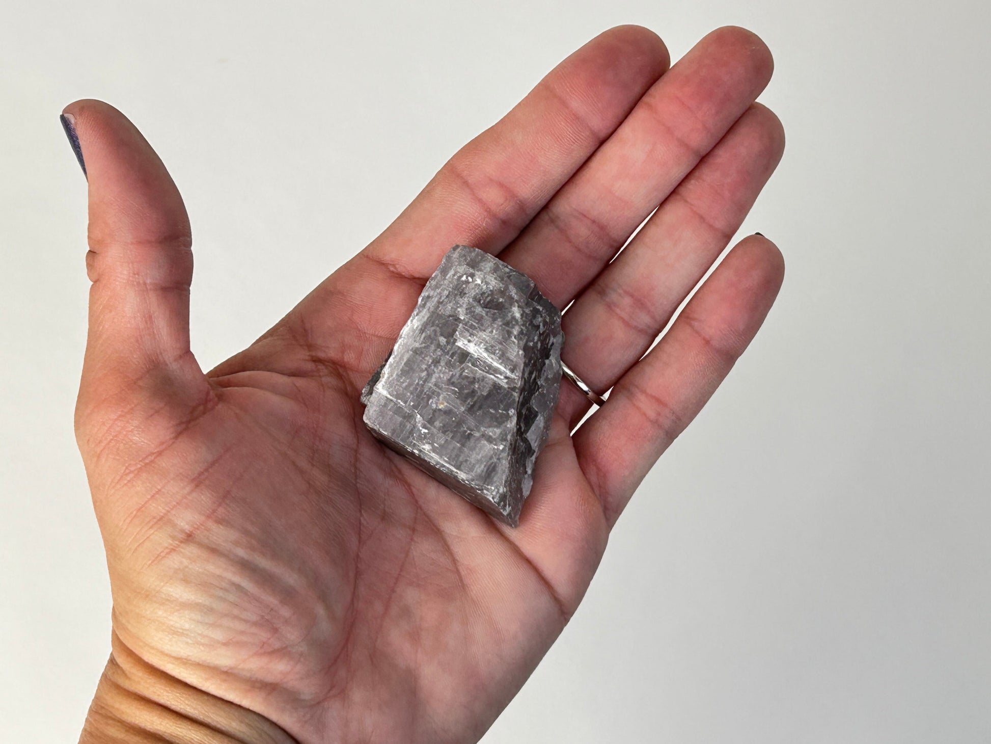A raw, rough gray metallic chunk of Soothing Purple Anhydrite Stone held in a person's open palm against a plain light background.