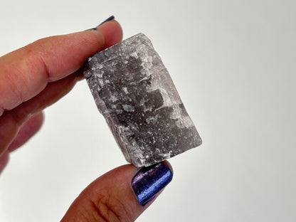 A hand holding a raw, rough rectangular translucent Soothing Purple Anhydrite Stone with visible internal fractures and natural edges against a plain white background.