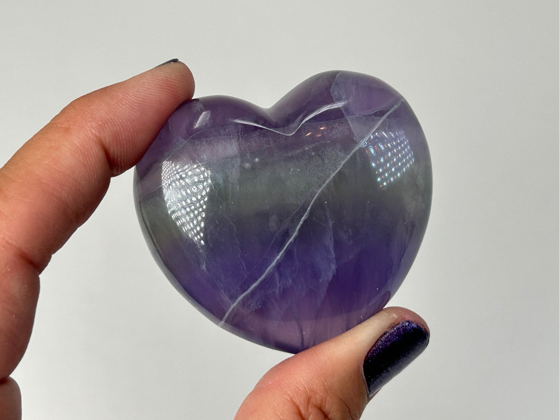 A polished purple heart-shaped Striped Fluorite Compassionate Heart Crystal held between a person's fingers against a plain light gray background.