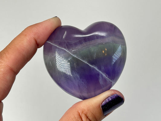 A hand holding a polished purple heart-shaped Striped Fluorite Compassionate Heart Crystal with natural white veining, set against a plain light gray background.