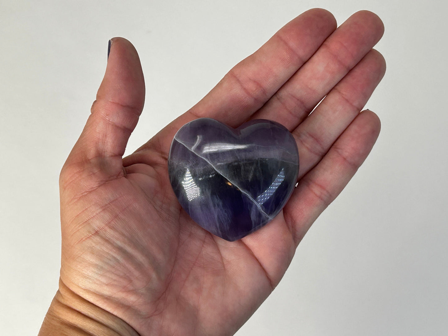 A polished heart-shaped purple Striped Fluorite Compassionate Heart Crystal held in a person's open palm against a plain light background.