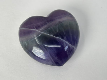 A polished heart-shaped Striped Fluorite Compassionate Heart Crystal with natural purple, blue, and white veining, photographed on a neutral light gray background.