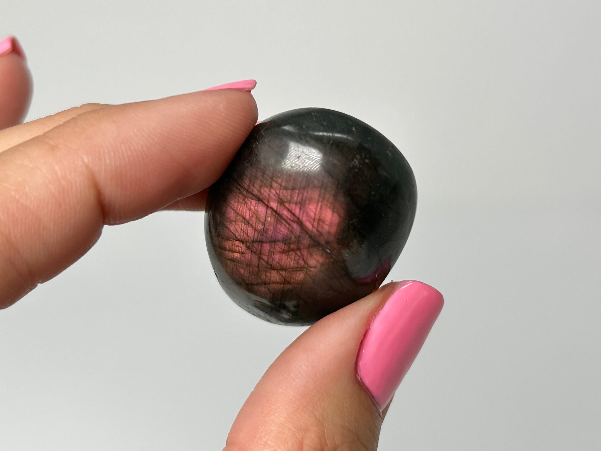 Person holding a dark Labradorite palm stone with pinkish-red streaks between their fingers against a plain background