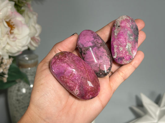 Three polished Cobalto Calcite reiki energized healing crystal Palm Stones for meditation energy balance and spiritual wellness held in a person's hand, with a blurred background suggesting a home altar setting with beautiful decor