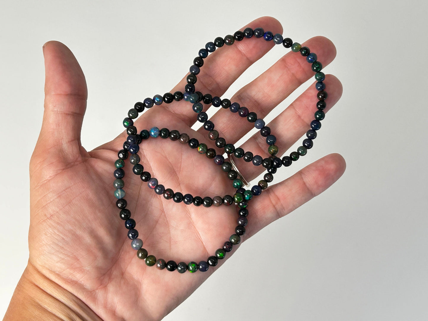 Hand holding a string of black and dark green/blue beaded opal bracelets against a plain background