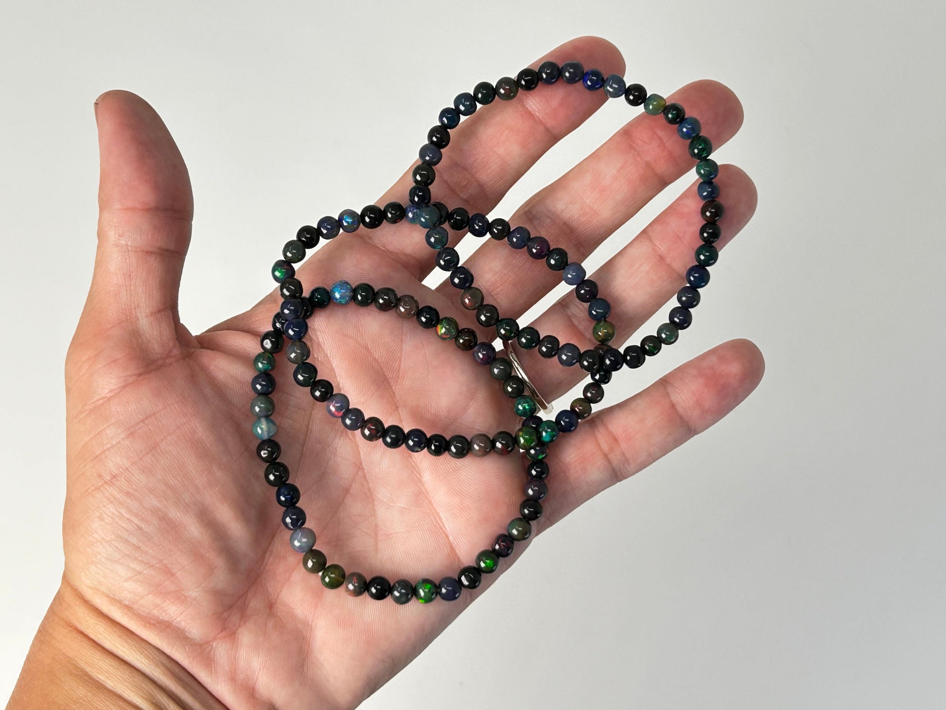 Hand holding a string of black and dark green/blue beaded opal bracelets against a plain background