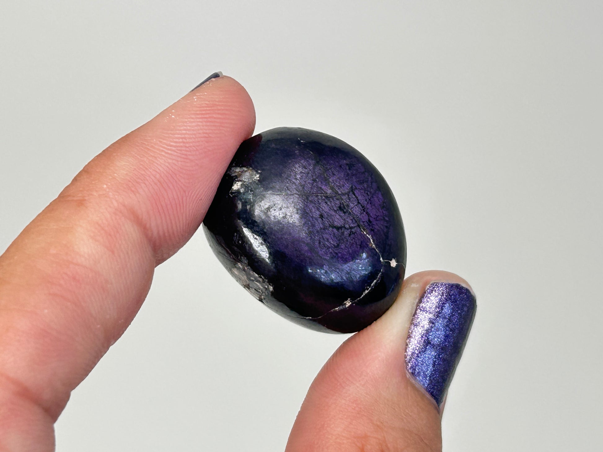 A polished dark purple Transformative Tumbled Purpurite Crystal held between two fingers against a plain light gray background.