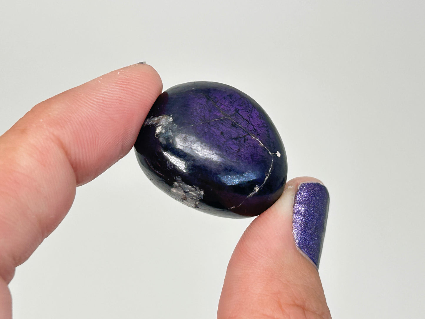 A hand holding a polished Transformative Tumbled Purpurite Crystal, showing its deep violet-blue color with natural white veining, against a plain light gray background.