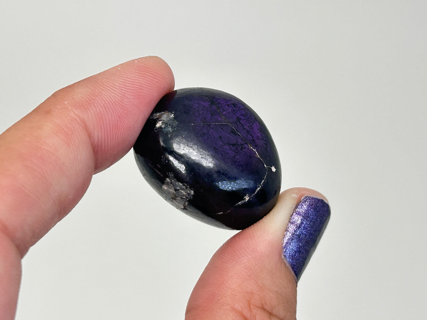 A polished dark purple and black Transformative Tumbled Purpurite Crystal  held between two fingers against a plain light gray background.