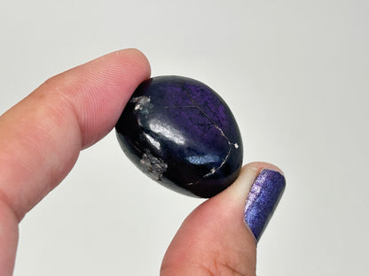 A polished dark purple and black Transformative Tumbled Purpurite Crystal  held between two fingers against a plain light gray background.