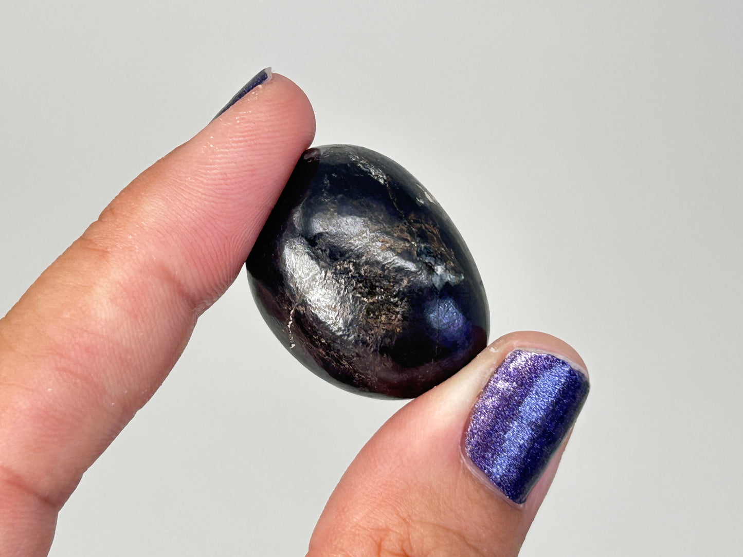 A hand with purple glitter nail polish holds a polished black Transformative Tumbled Purpurite Crystal with natural metallic silver and gray veins against a plain light gray background.