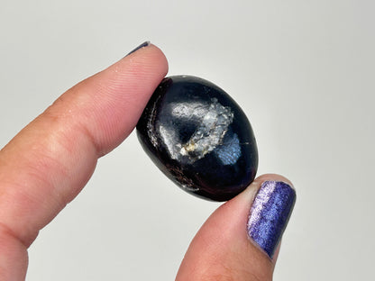 A polished dark blue Transformative Tumbled Purpurite Crystal held between two fingers against a plain light gray background.