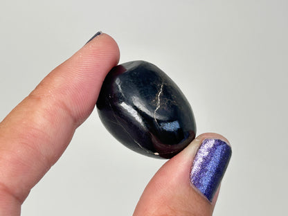 A polished dark Transformative Tumbled Purpurite Crystal held between two fingers against a plain light gray background.