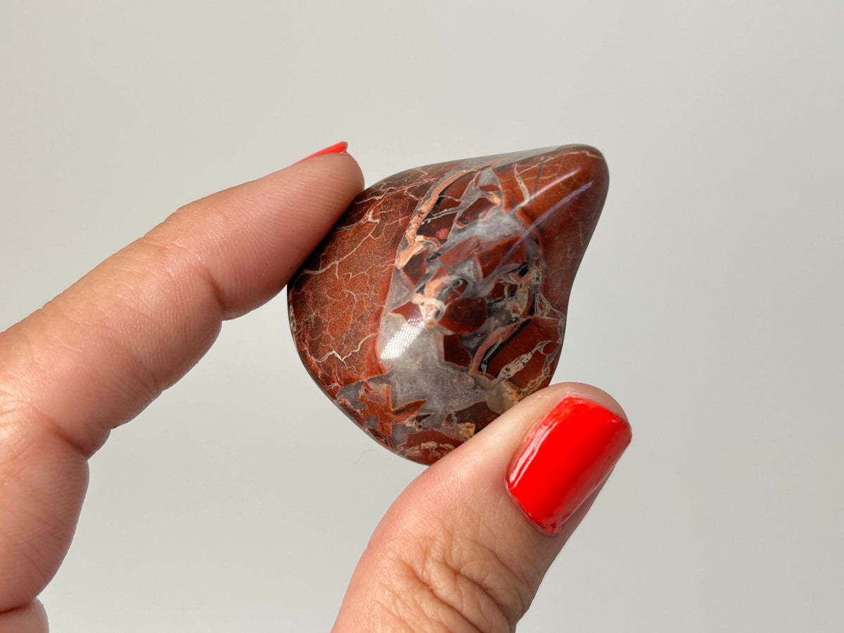 A polished red and brown Tumbled Brecciated Jasper crystal with natural white and gray veining, held between two fingers against a plain light gray background.