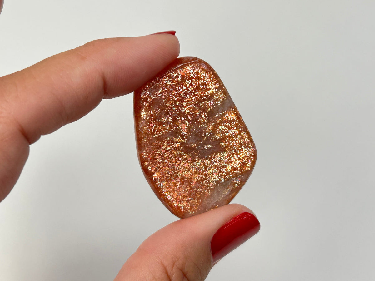 A hand with red nail polish holds a Tumbled Confetti Sunstone crystal  covered in fine rose-gold and copper glitter against a plain light gray background.
