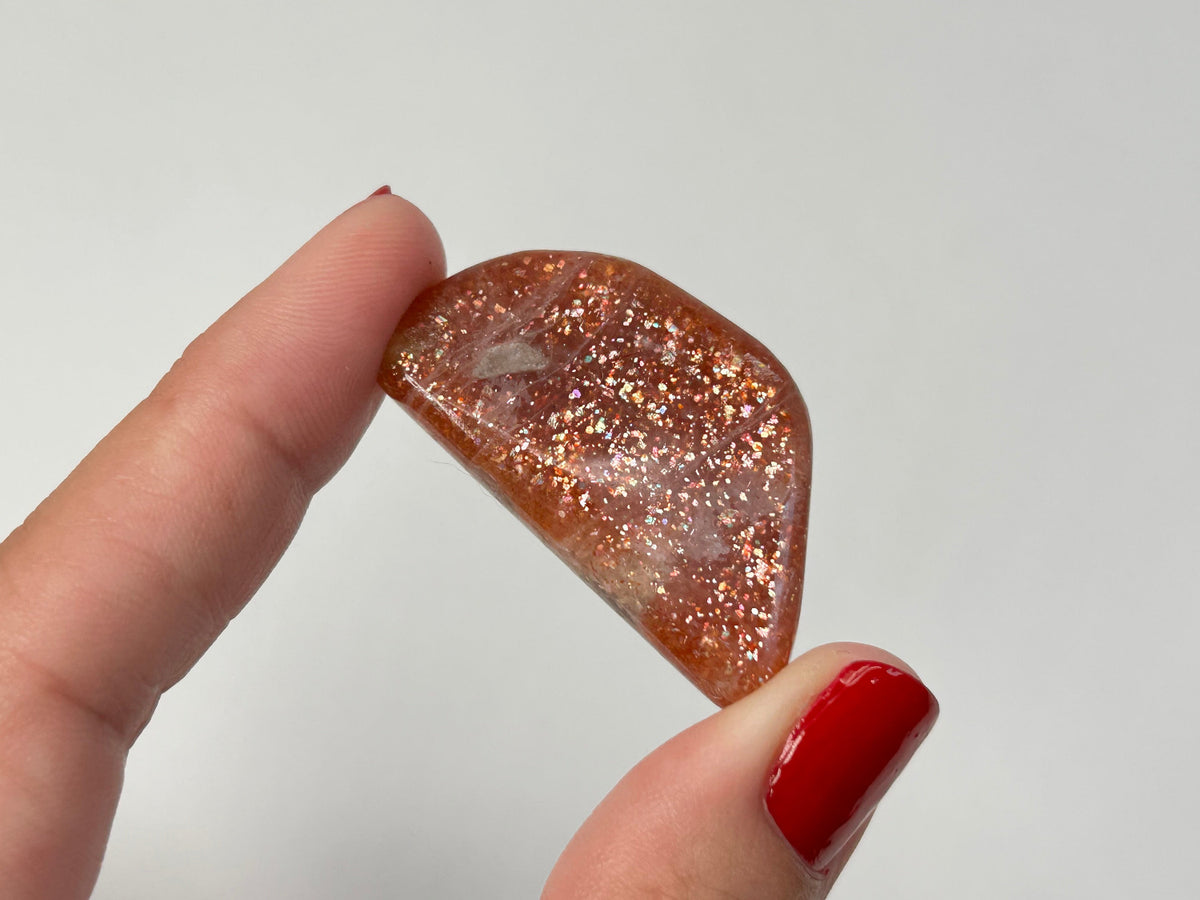 A hand with red nail polish holds a polished,Tumbled Confetti Sunstone for healing and peace-of-mind filled with shimmering copper and gold glitter against a plain light gray background.
