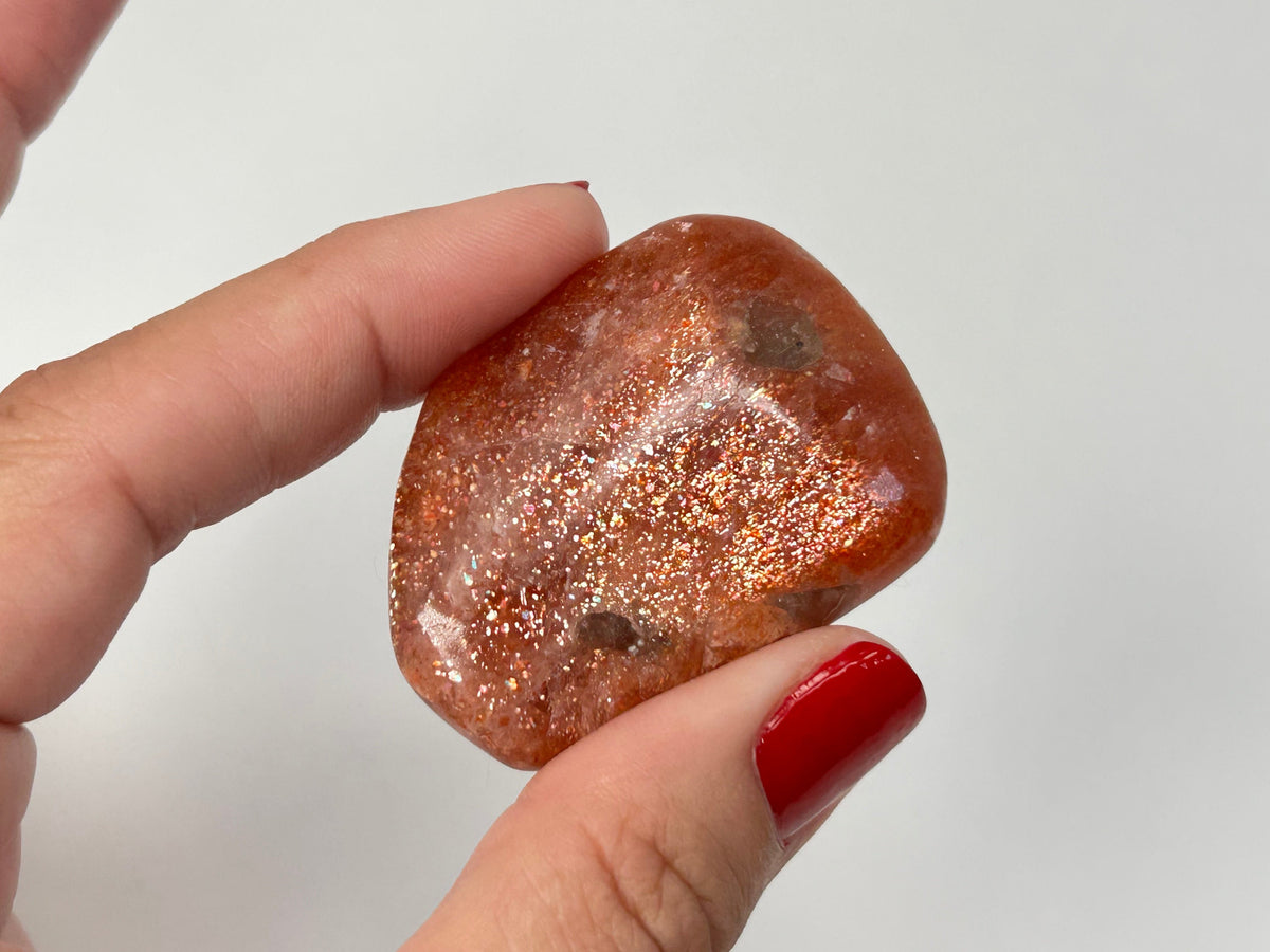 A hand with red painted fingernails holds a polished Tumbled Confetti Sunstone gemstone with a coppery orange-brown body and fine glitter, set against a plain light gray background.