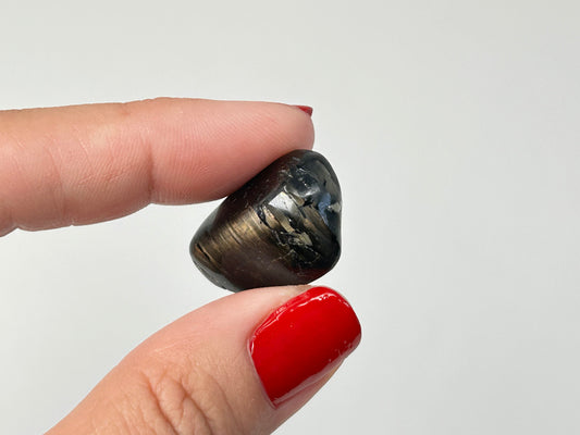 A polished dark metallic Tumbled Nuummite gemstone held between two fingers with red nail polish against a plain light gray background.