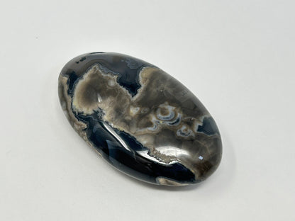 A polished oval Volcano Agate Crystal Healing Palm Stone with natural marbled patterns of black, gray, and white, displayed on a clean white background.
