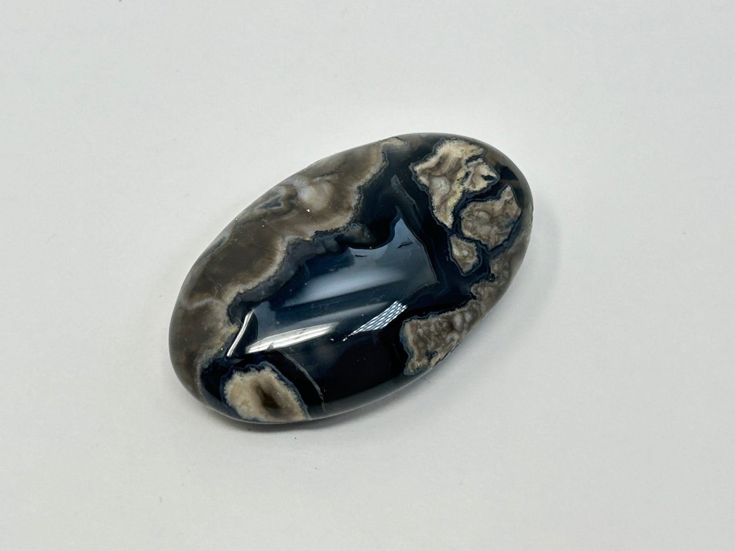 A polished oval Volcano Agate Crystal Healing Palm Stone featuring a marbled pattern of glossy black, dark gray, and beige veins, photographed on a neutral light gray background.