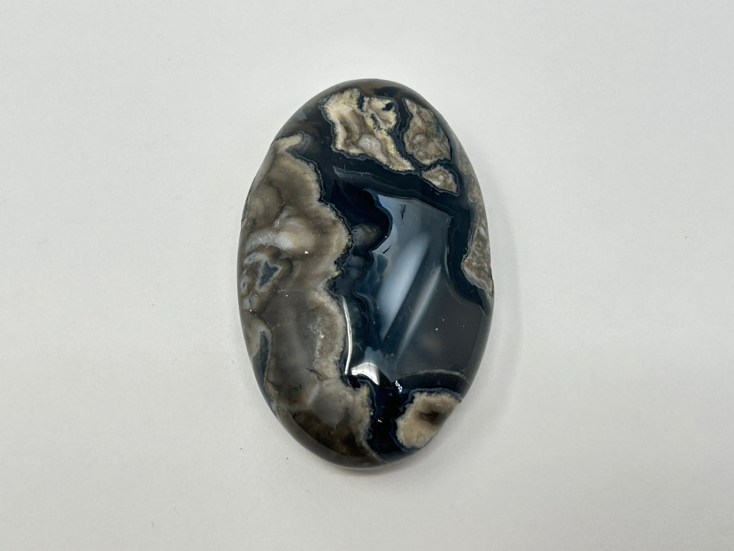 A polished oval Volcano Agate Crystal Healing Palm Stone for creativity mindfulness and curiosity featuring a marbled pattern of black, dark gray, and beige, photographed on a plain light background.
