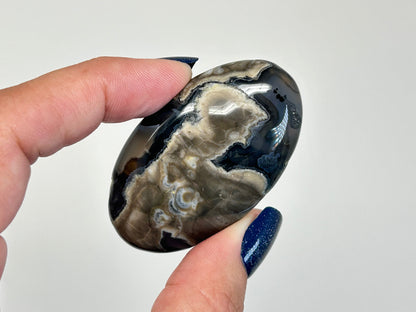 A hand with dark blue glitter nail polish holds a polished, oval-shaped Volcano Agate Crystal Healing Palm Stone featuring natural marbled patterns of black, gray, and beige, against a plain light gray background.