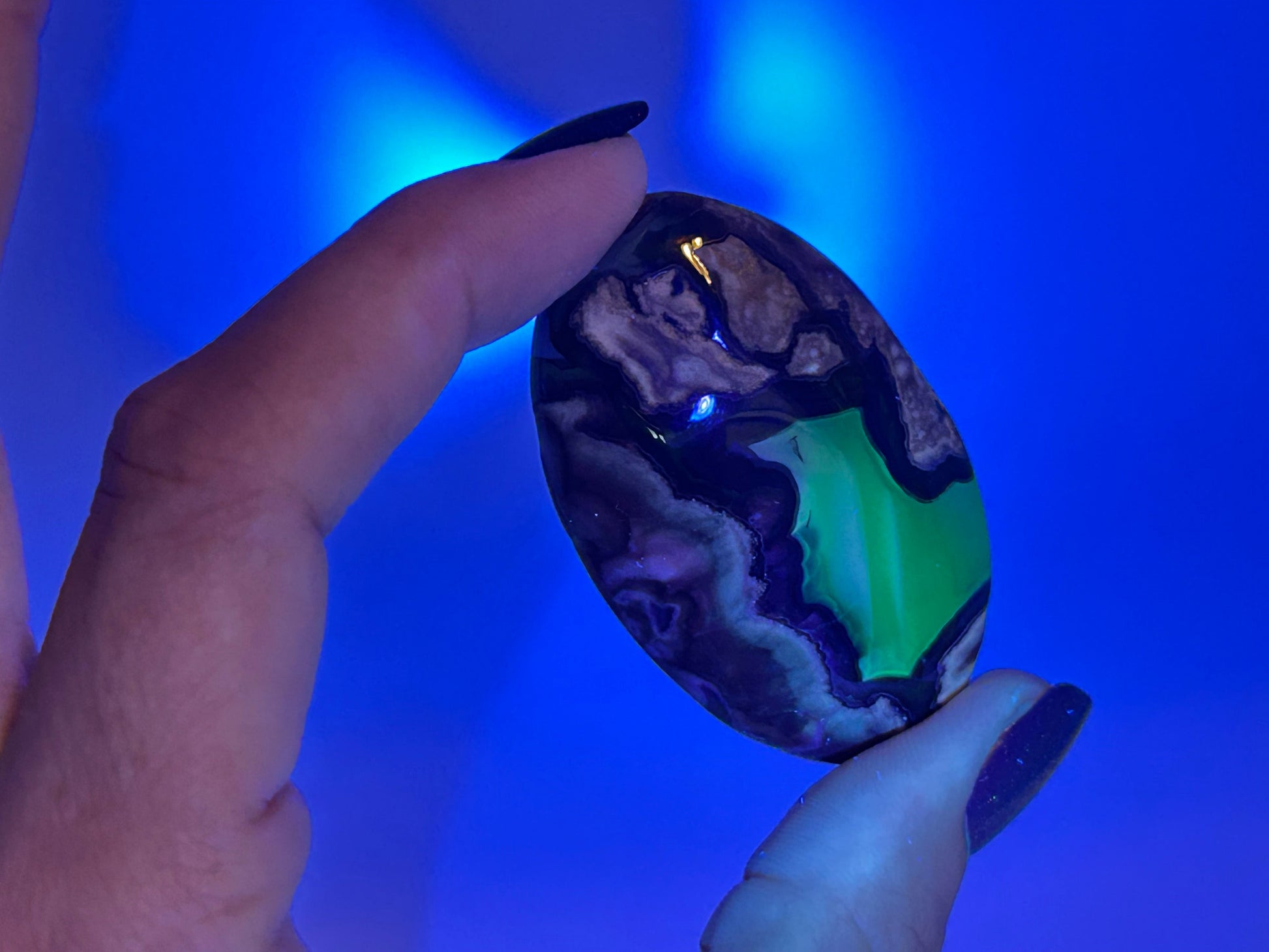A hand with dark glittery fingernails holds a polished oval Volcano Agate Crystal Healing Palm Stone under blue light, which highlights its natural marbled pattern of black, purple, brown, and bright neon green.