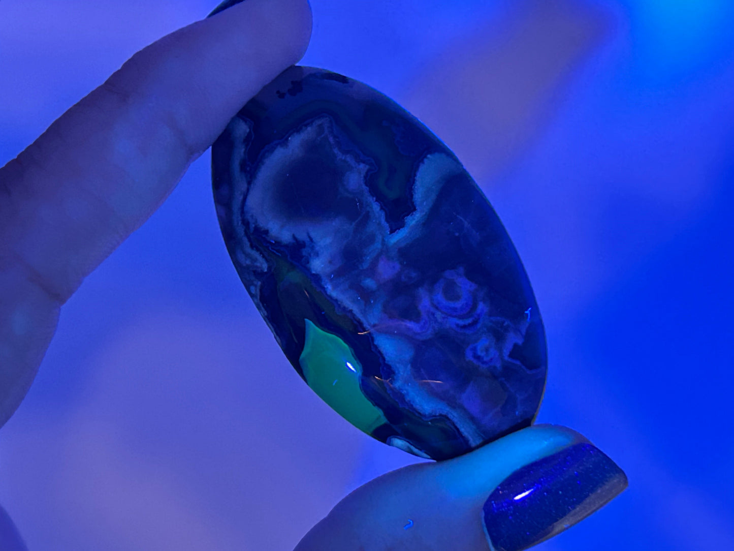 A hand holding a polished oval Volcano Agate Crystal Healing Palm Stone under blue light, showing deep blue, black, white, and green marbled patterns.