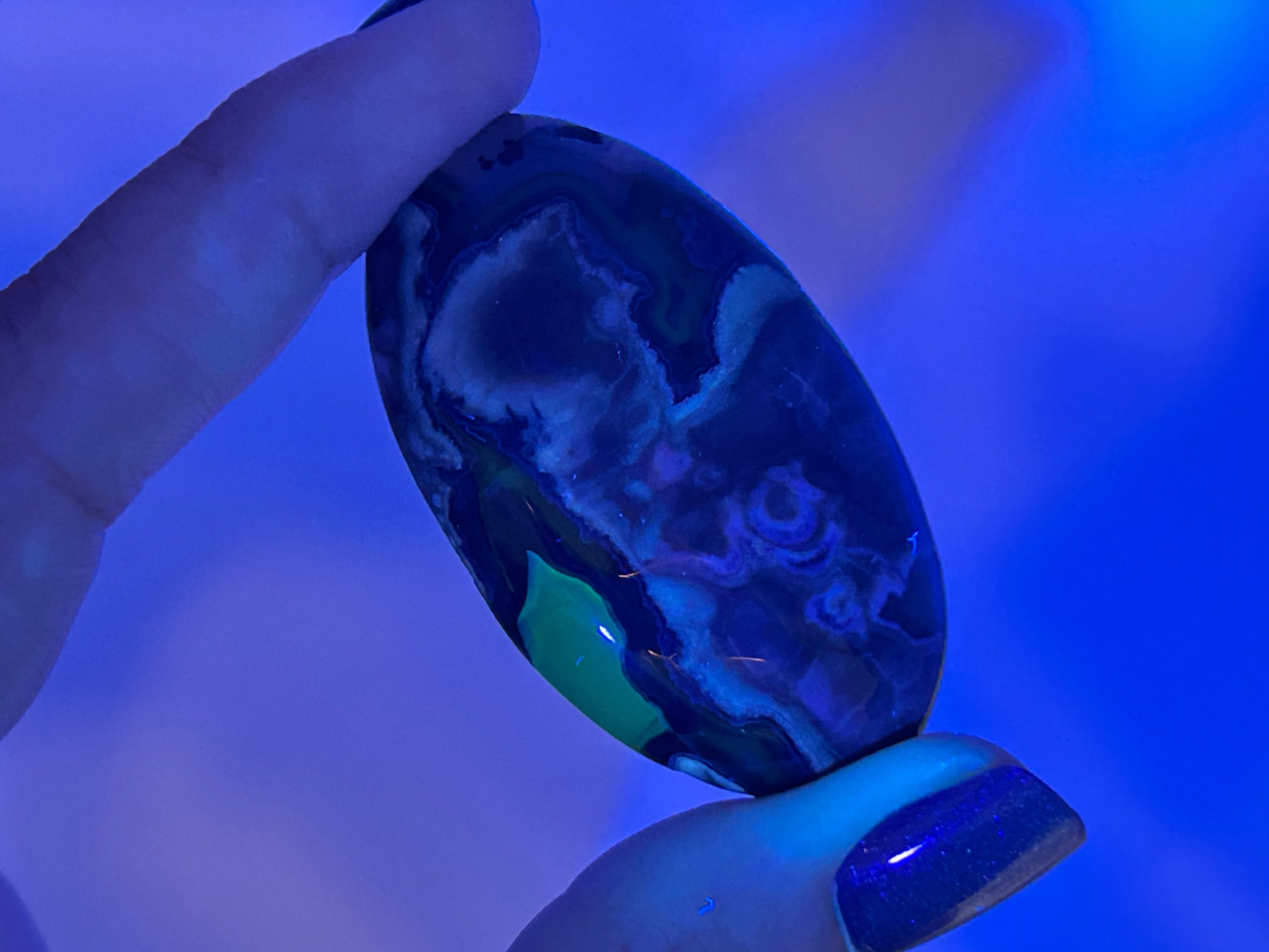 A hand holding a polished oval Volcano Agate Crystal Healing Palm Stone under blue light, showing deep blue, black, white, and green marbled patterns.