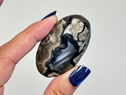 A hand with dark blue glitter nail polish holds a polished, oval-shaped Volcano Agate Crystal Healing Palm Stone with swirling patterns of black, gray, and beige, against a plain light gray background.