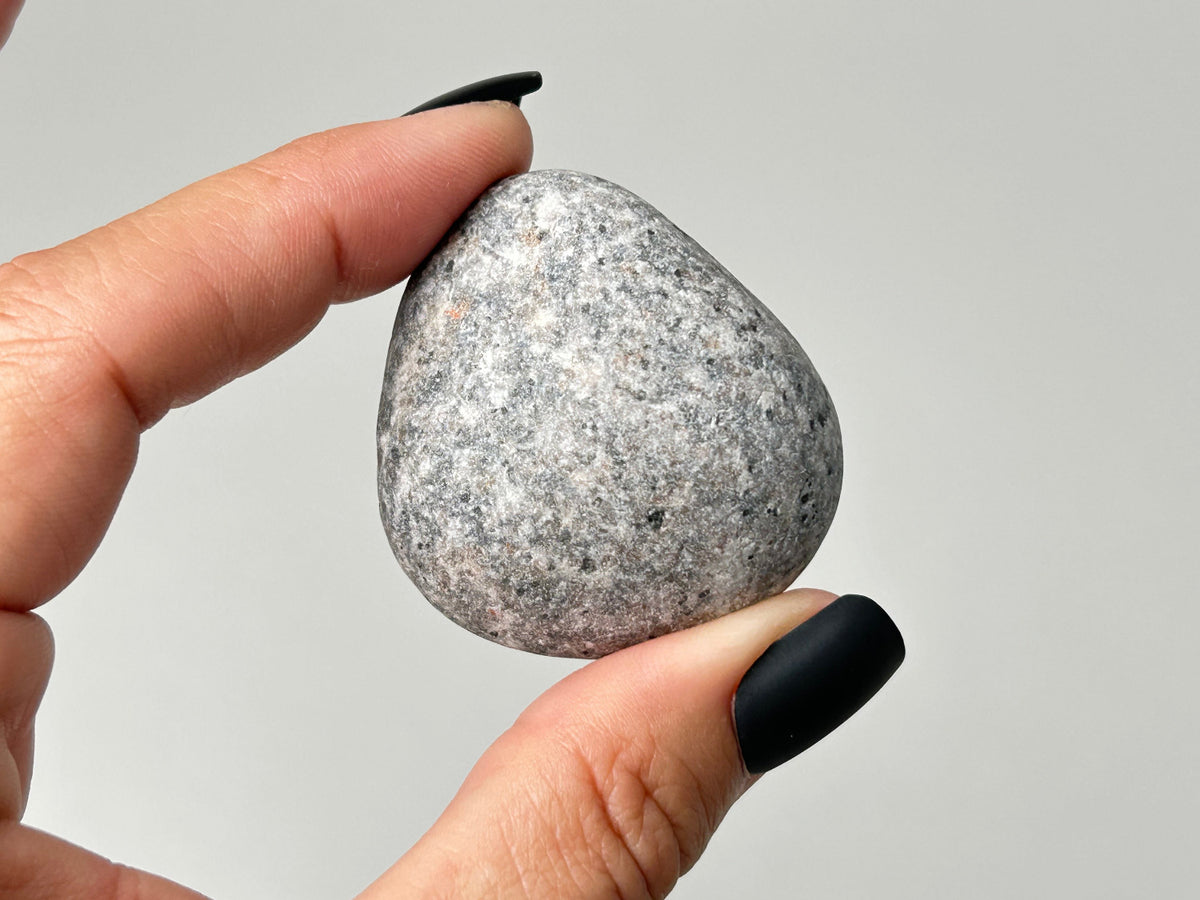 A hand with black manicured nails holds a smooth, rounded gray speckled Yooperlite fluorescent Pebble against a plain light gray background.