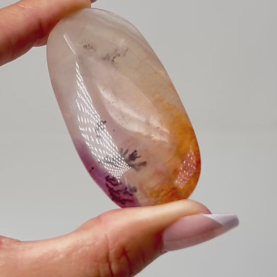 Hand holding a Polished, oval-shaped light-brown Dendritic Fluorite mineral palm stone with brown and orange colored markings reminiscent of a landscape healing crystal gemstone for meditation and energy work with white background.