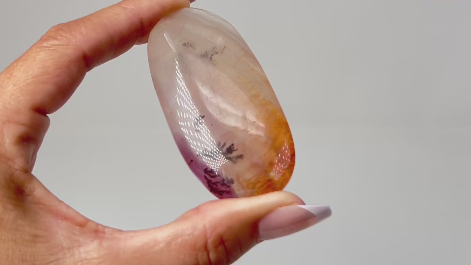 Hand holding a Polished, oval-shaped light-brown Dendritic Fluorite mineral palm stone with brown and orange colored markings reminiscent of a landscape healing crystal gemstone for meditation and energy work with white background.