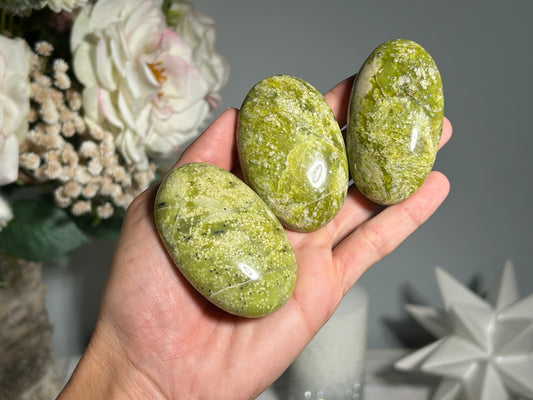Three polished green serpentine palm stones held in a person's hand, with a blurred background suggesting indoor setting